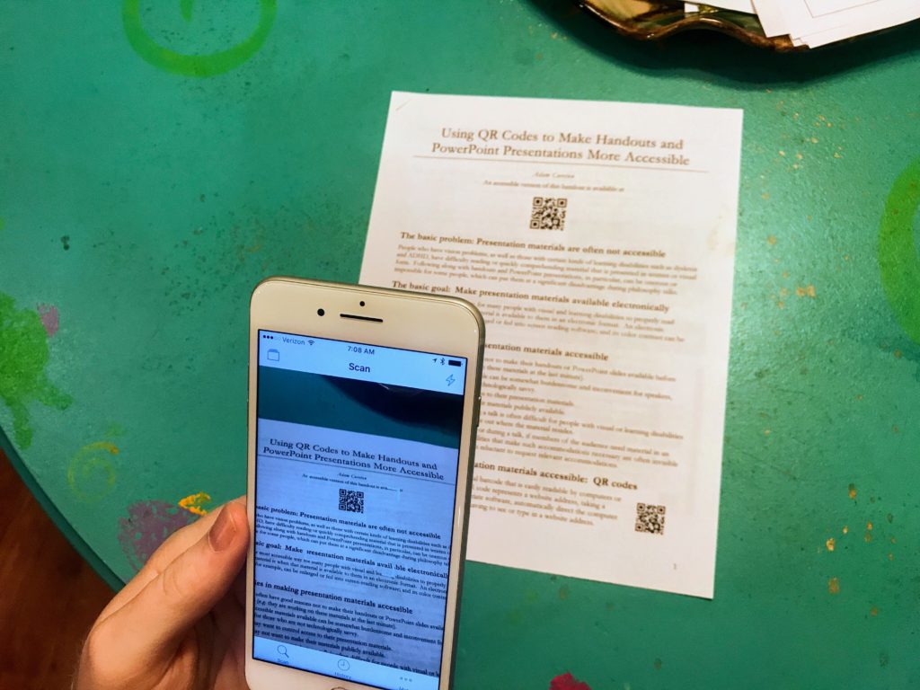 Shows an Apple iPhone pointed at a document with a QR code on it. The phone has a QR code reader app open and shows an image of the document with the QR code in the middle.