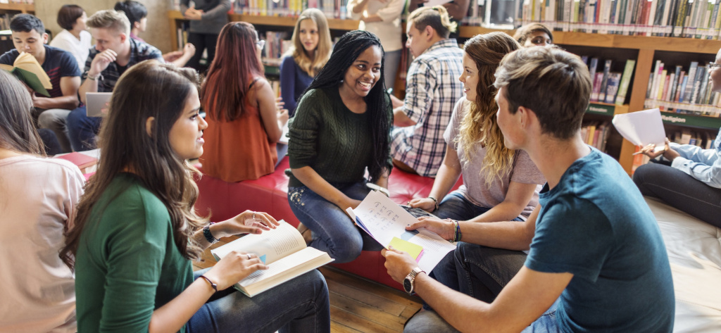 A group of students sit and talk together in a library. The atmosphere is warm and collaborative.